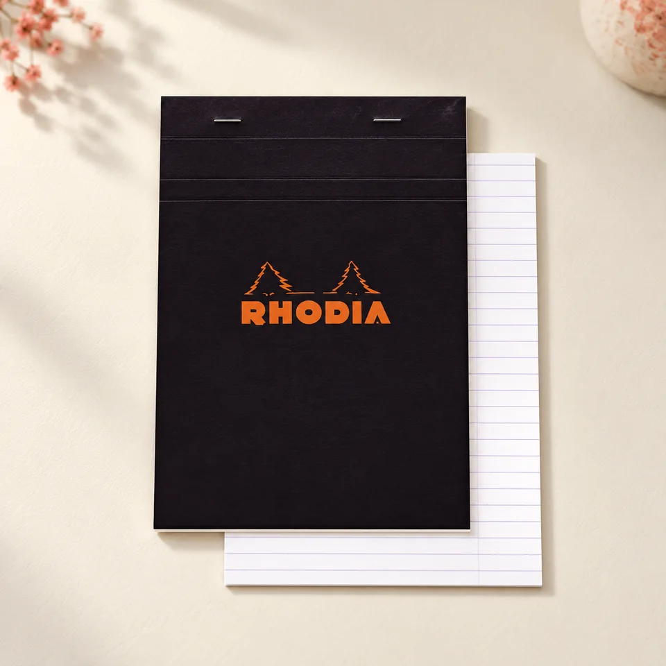 Rhodia — bloc agrafé A5 noir de 80 feuillets de Rhodia, mise en situation dans un atelier de papier écru et corail